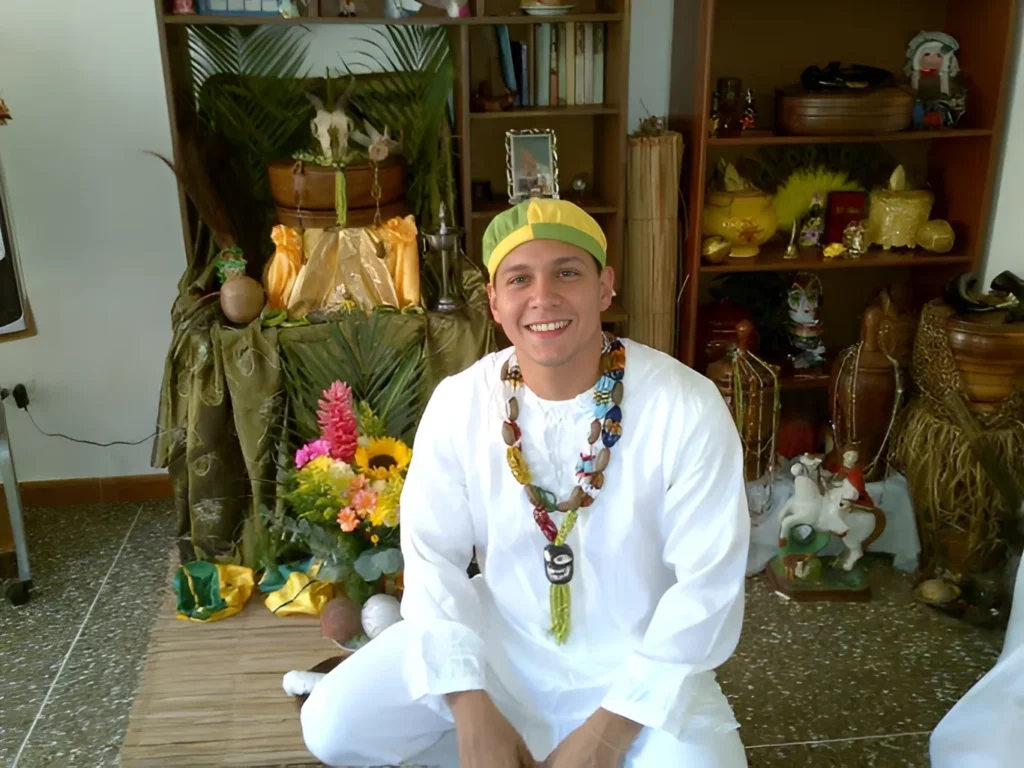 Aniversario de Ifa Babalawo de Ifá y creador de Ile ifa Imo, sonríe radiante en su primer aniversario de Ifá, frente a un altar con ofrendas y objetos sagrados